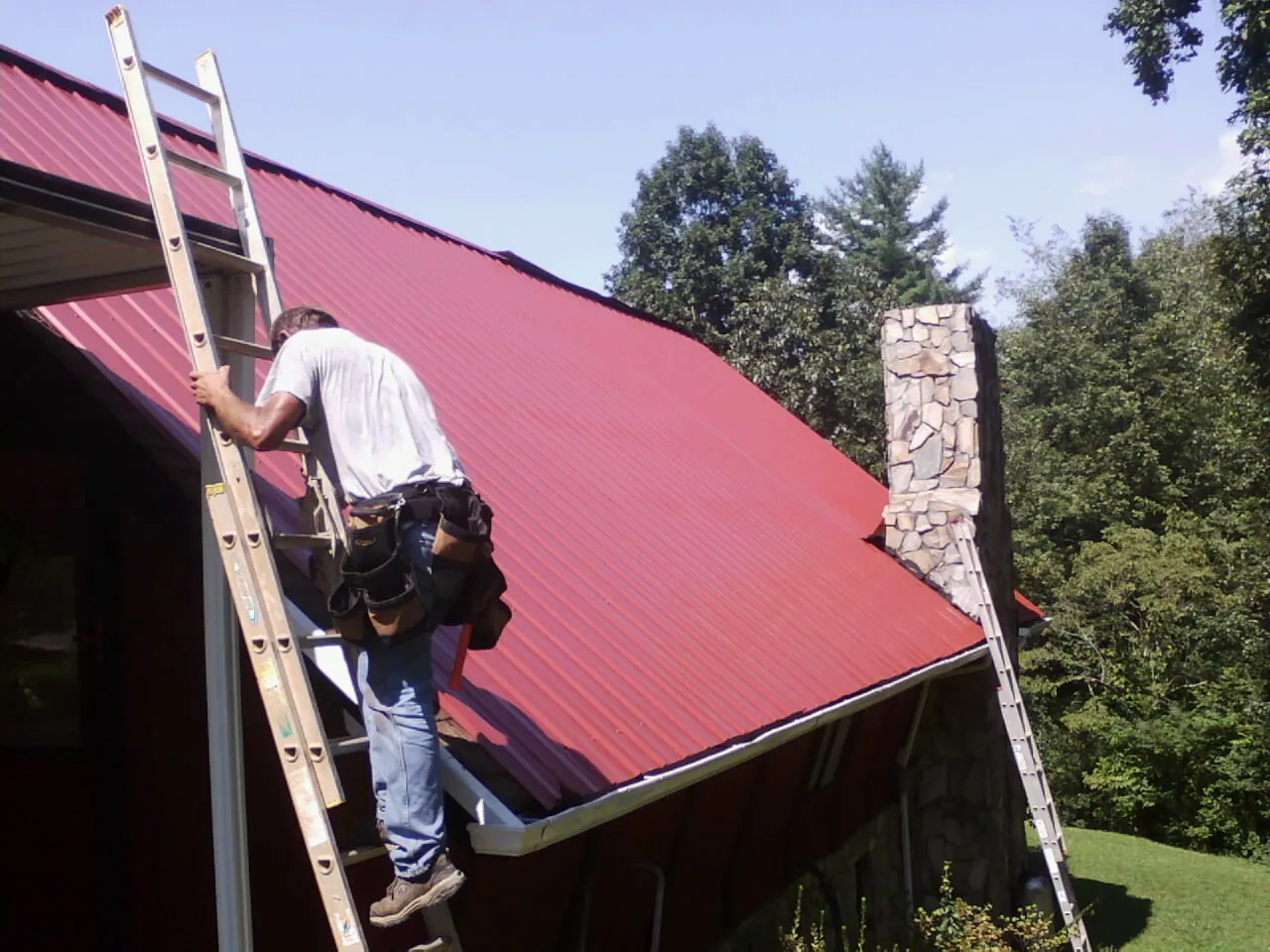 Roofer on a ladder beside a red metal roof and stone chimney — Soffit & Fascia Repair by Roofing Brothers in Parker Ford